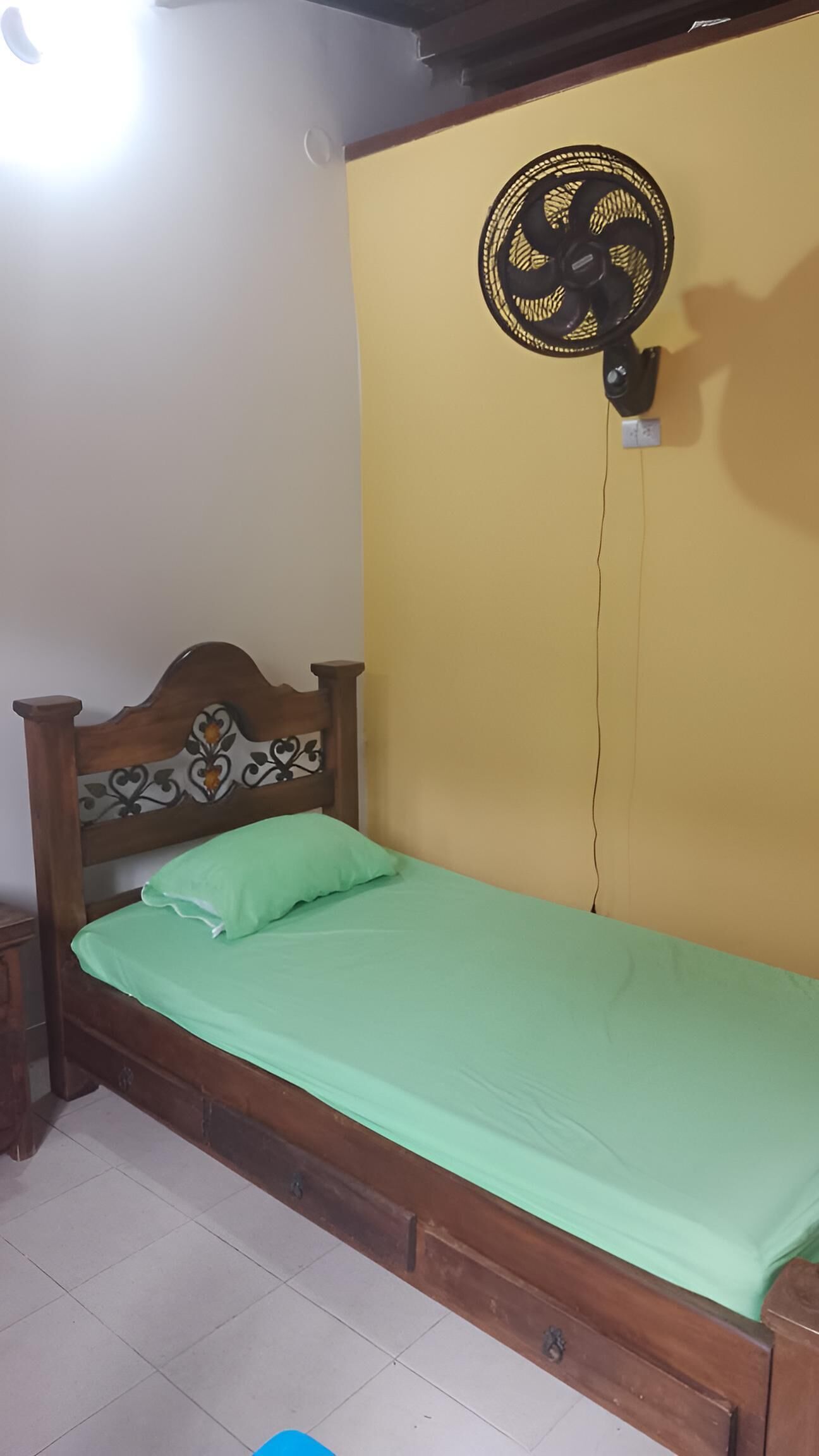 Uma cama de solteiro com cabeceira de madeira e colchão verde, um ventilador fixado na parede e piso ladrilhado.