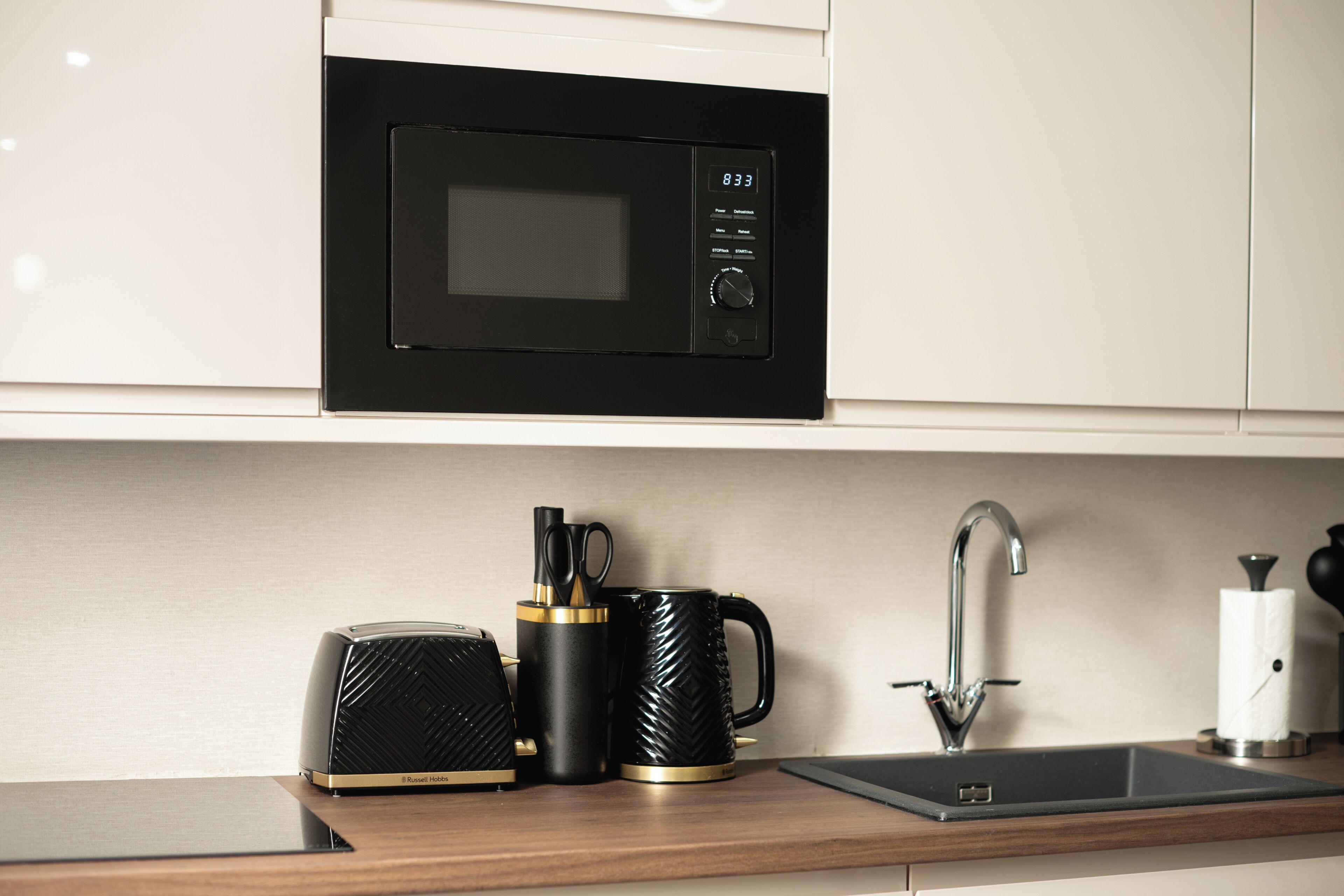 Una cocina con microondas negro, tostadora, cafetera, fregadero y dispensador de toallas de papel.