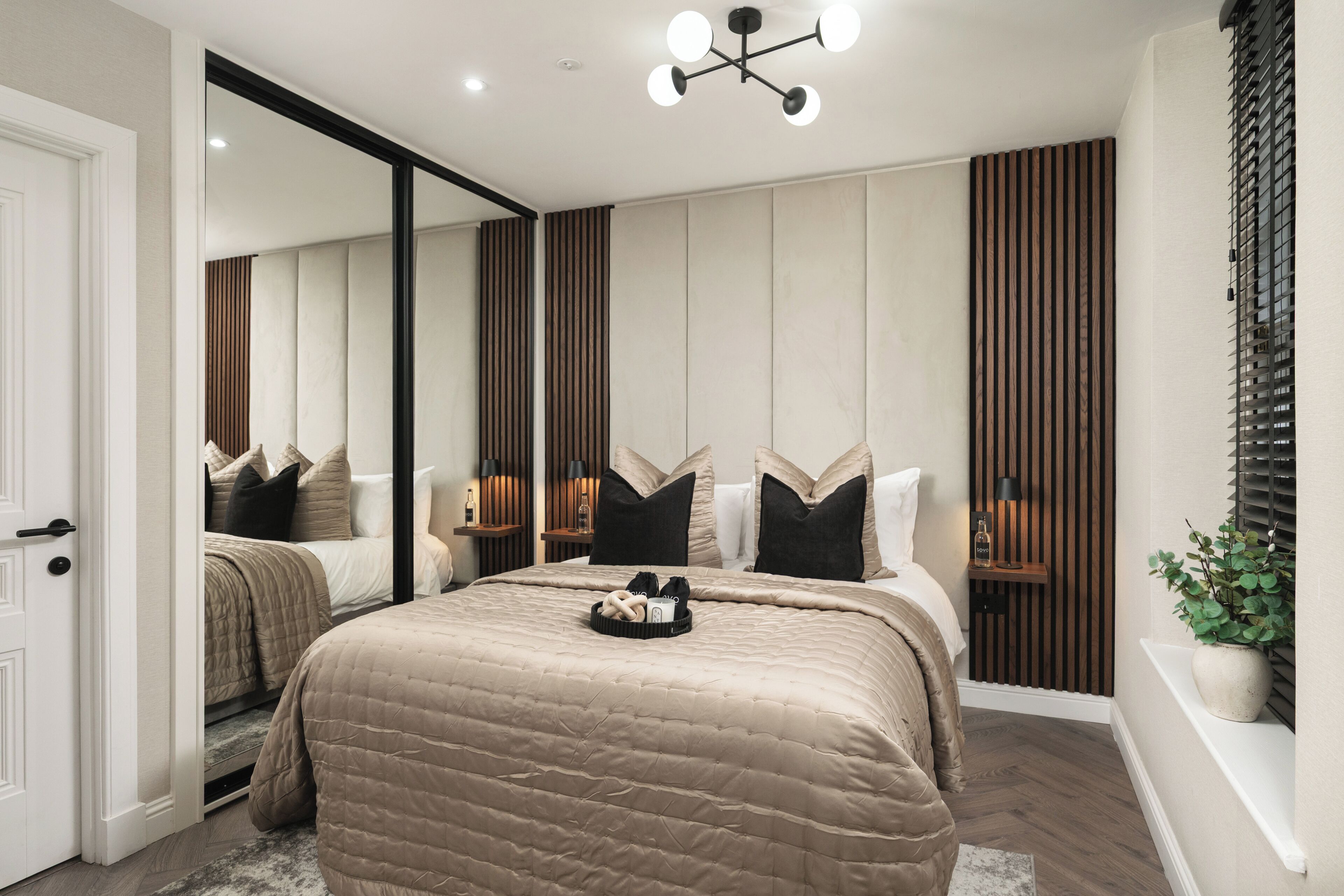 Quarto moderno com uma cama grande, um espelho, uma mesa de cabeceira com abajur e uma planta em vaso.