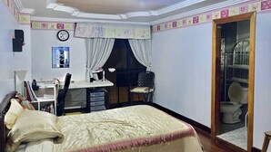 Ein Schlafzimmer mit Bett, Schreibtisch, Stuhl und einem durch eine offene Tür einsehbarer Badezimmer.