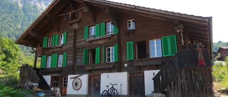 A wooden chalet with green shutters, a bicycle, and a mountain backdrop.