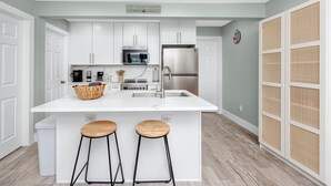 A modern kitchen with white cabinetry, a stainless steel refrigerator, a microwave, and a coffee maker. There are two wooden stools at the island. - Cottage Pool Across From the Beach (Madeira Beach)