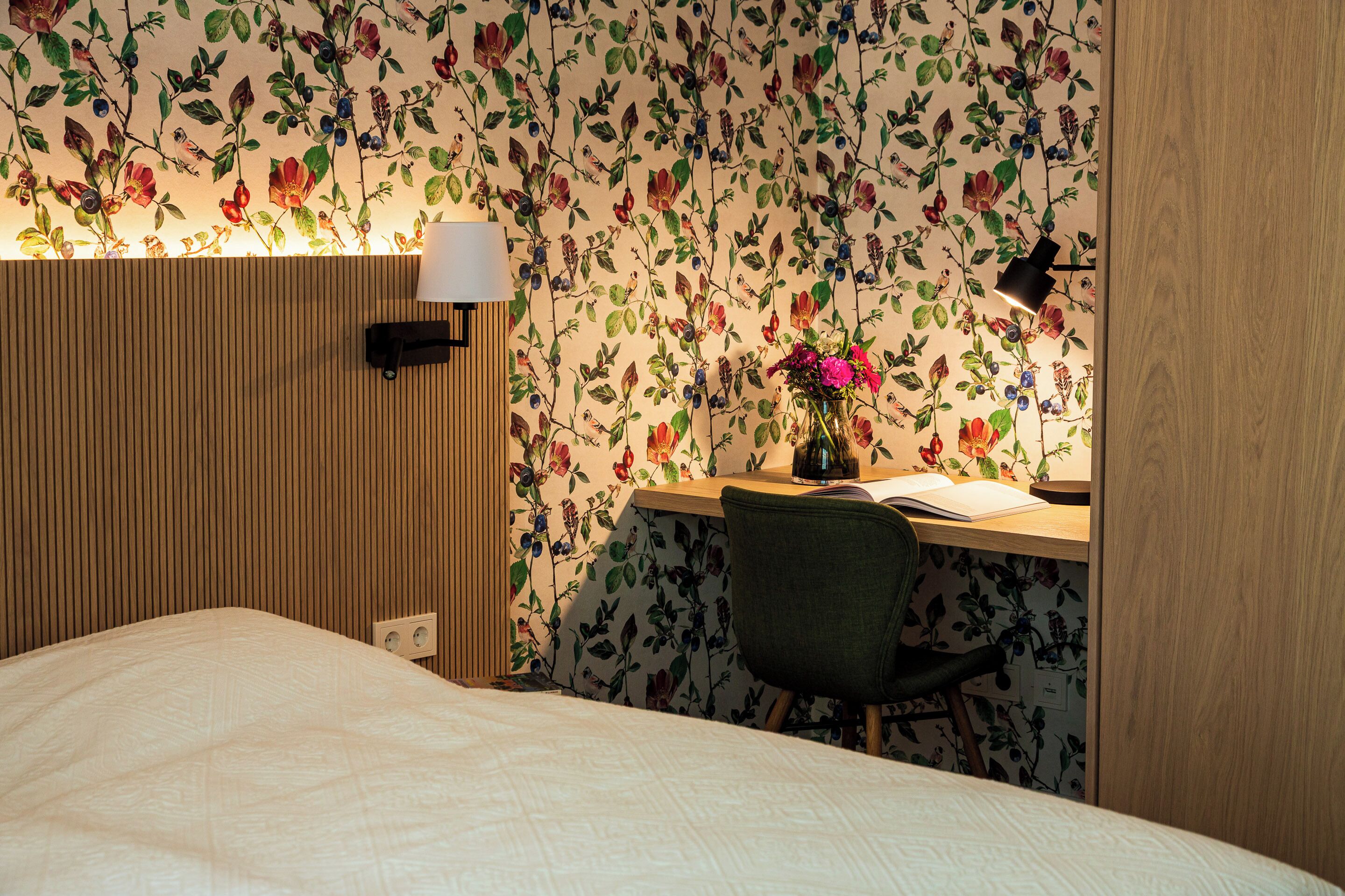 A bedroom with a bed, a desk with a chair, a lamp, and a wall with floral wallpaper.