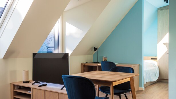 Una stanza con soffitto spiovente, una TV su un supporto in legno, una scrivania con una sedia e un letto con copripiumino bianco.