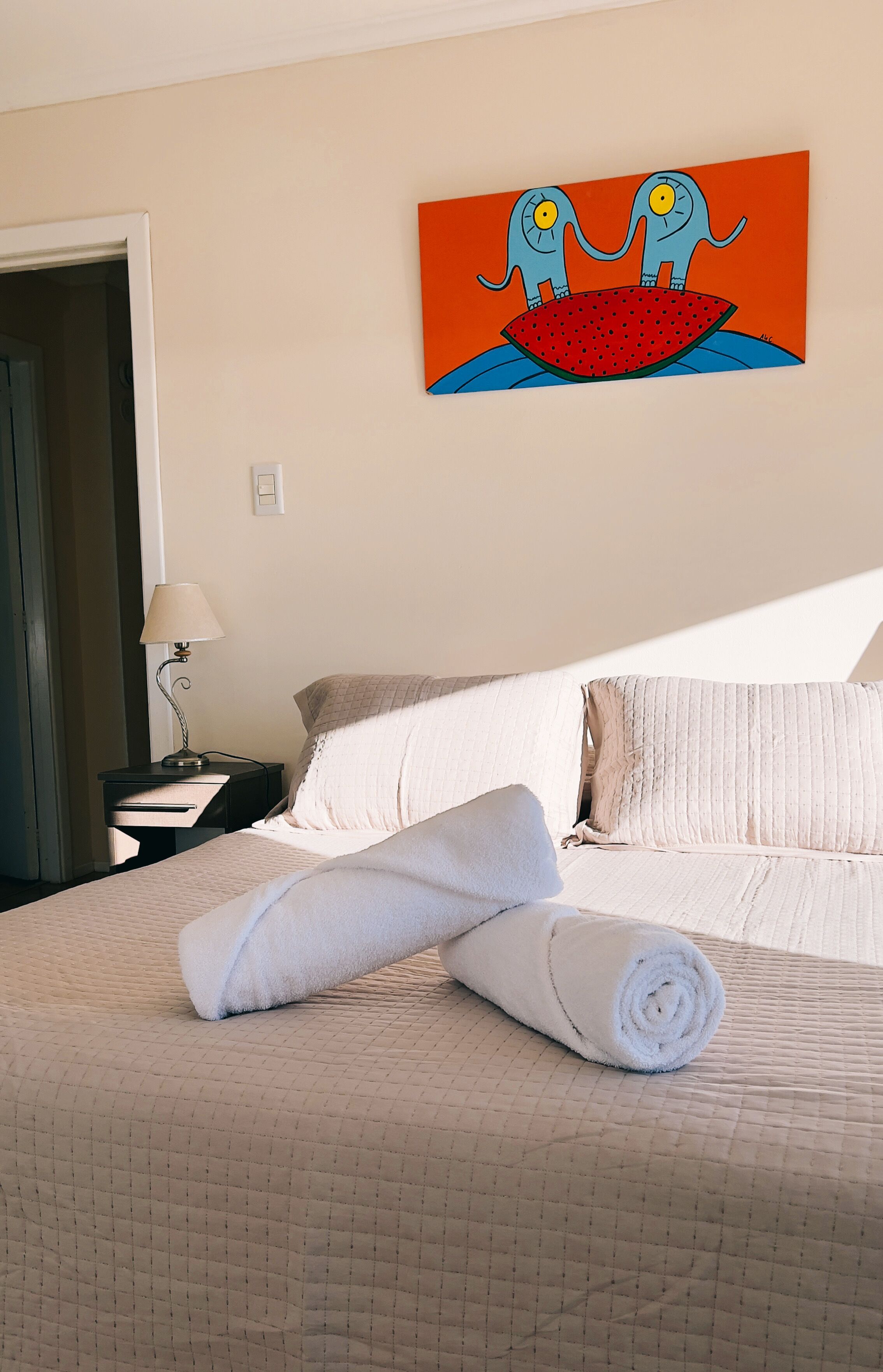 Quarto de hotel com cama, almofadas, toalha enrolada, mesa de cabeceira com abajur e uma pintura na parede.