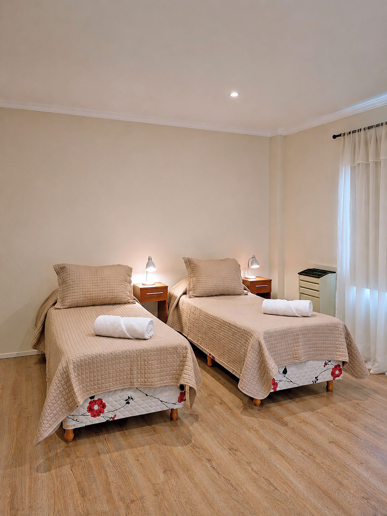 Two twin beds with matching headboards, bedside tables with lamps, and a window with sheer curtains.