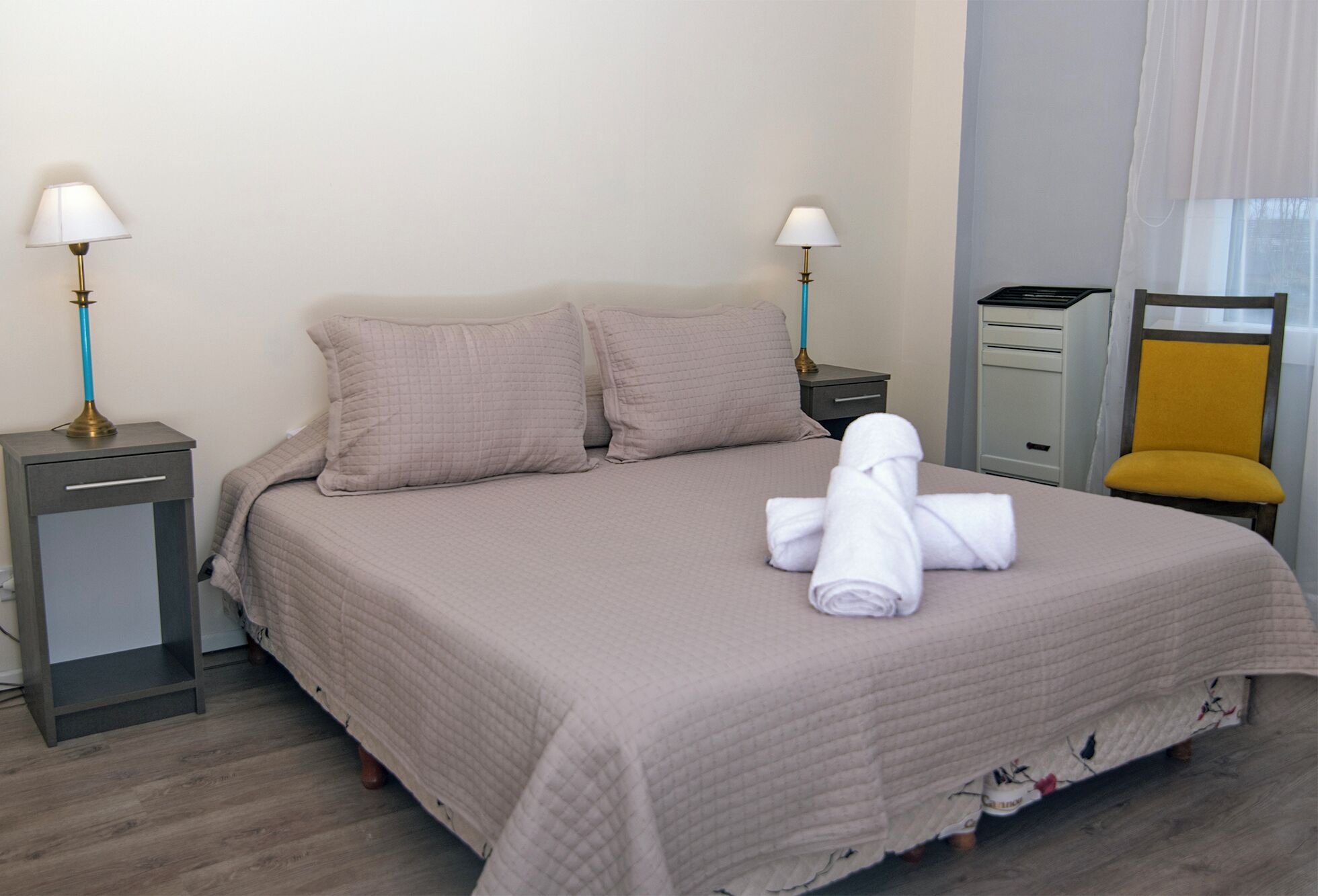 Una cama bien hecha con una colcha acolchada, dos almohadas, una toalla doblada, una mesita de noche con una lámpara, una silla y un aparador.