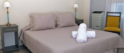 Una cama bien hecha con una colcha acolchada, dos almohadas, una toalla doblada, una mesita de noche con una lámpara, una silla y un aparador.