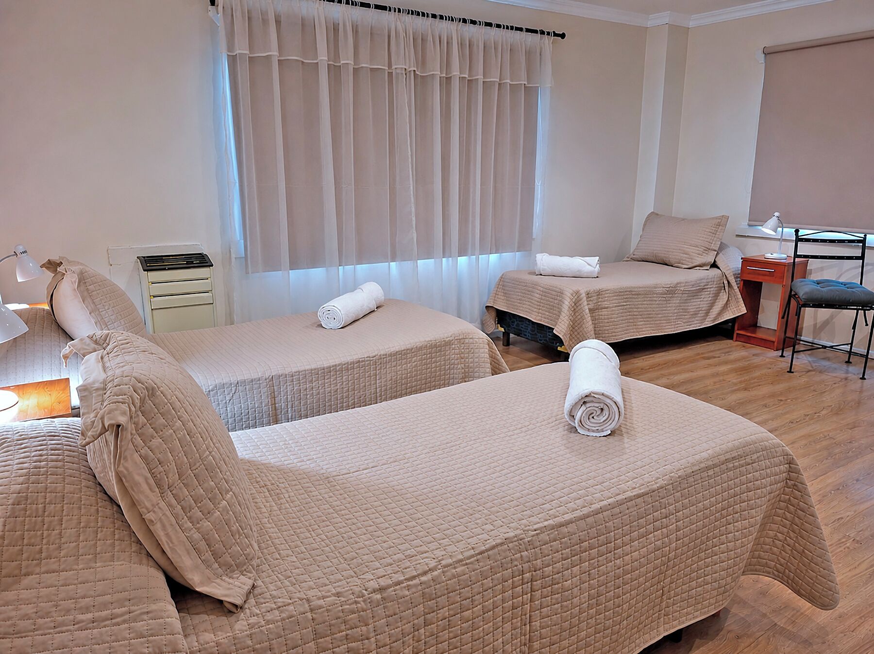 A hotel room with two single beds, a desk, a chair, a nightstand, and a window with curtains.