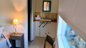 Ein kleines Zimmer mit einem Bett, einem Nachttisch mit Lampe, einem Sessel, einem Badezimmer mit Waschbecken und Spiegel sowie einem Fenster mit Blick nach außen.