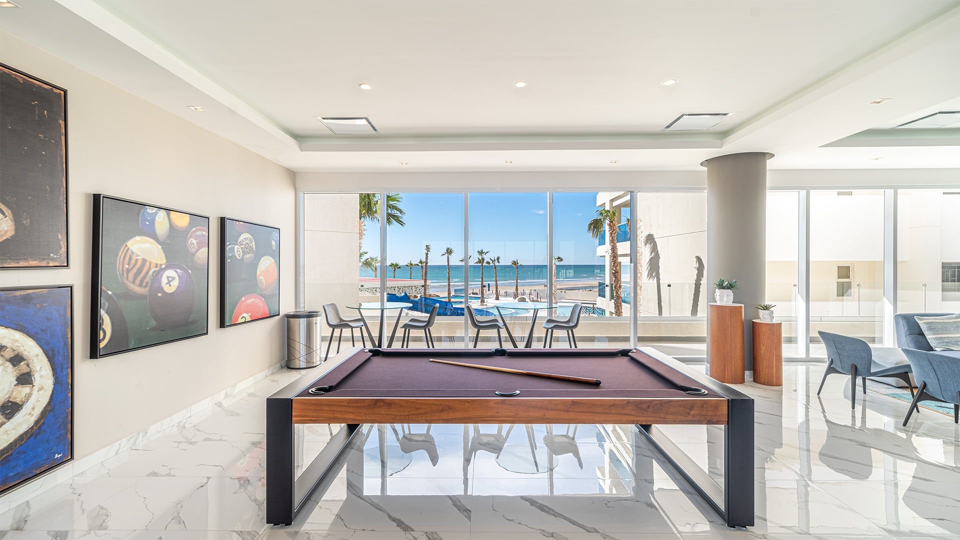 Mesa de billar con vista a la playa, obras de arte enmarcadas y muebles modernos.