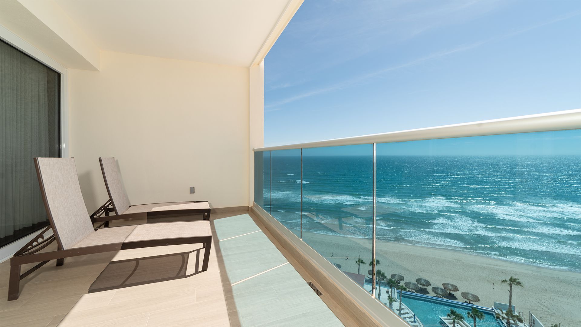 A balcony with a view of the ocean, a pool, and lounge chairs.