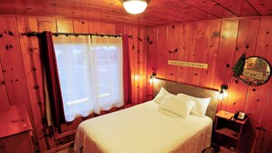 A wooden room with a bed, a nightstand, a wall-mounted light, and a window with curtains. - Kitchenette 1: Cozy Woodsy Suite w/  Kitchen – Your Adventure Basecamp (Florence)
