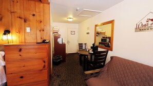 Hotellihuone, jossa on puinen lipasto, sänky, ruokapöytä tuoleineen, peili ja seinällä kyltti.