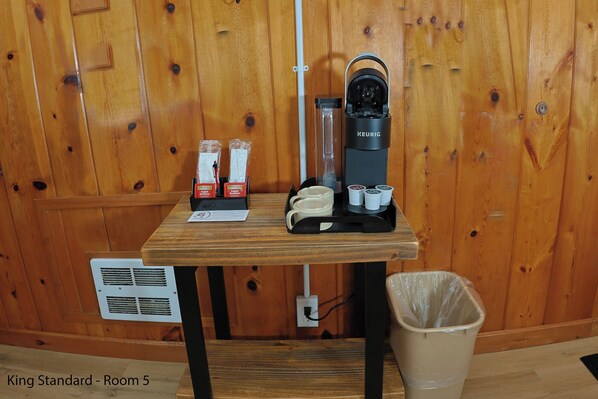 Una estación de café con una cafetera Keurig, vasos desechables y un cubo de basura en una habitación con paredes de madera.
