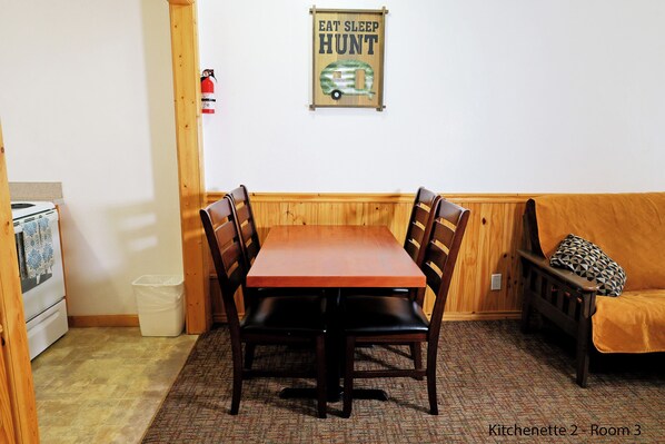 A dining area with a wooden table and chairs, a fire extinguisher, a whiteboard, and a couch with a patterned pillow.