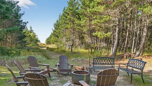A forested area with Adirondack chairs and a fire pit.