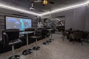 Una habitación moderna con una pantalla grande, asientos cómodos, un ventilador de techo y un mural original en la pared.