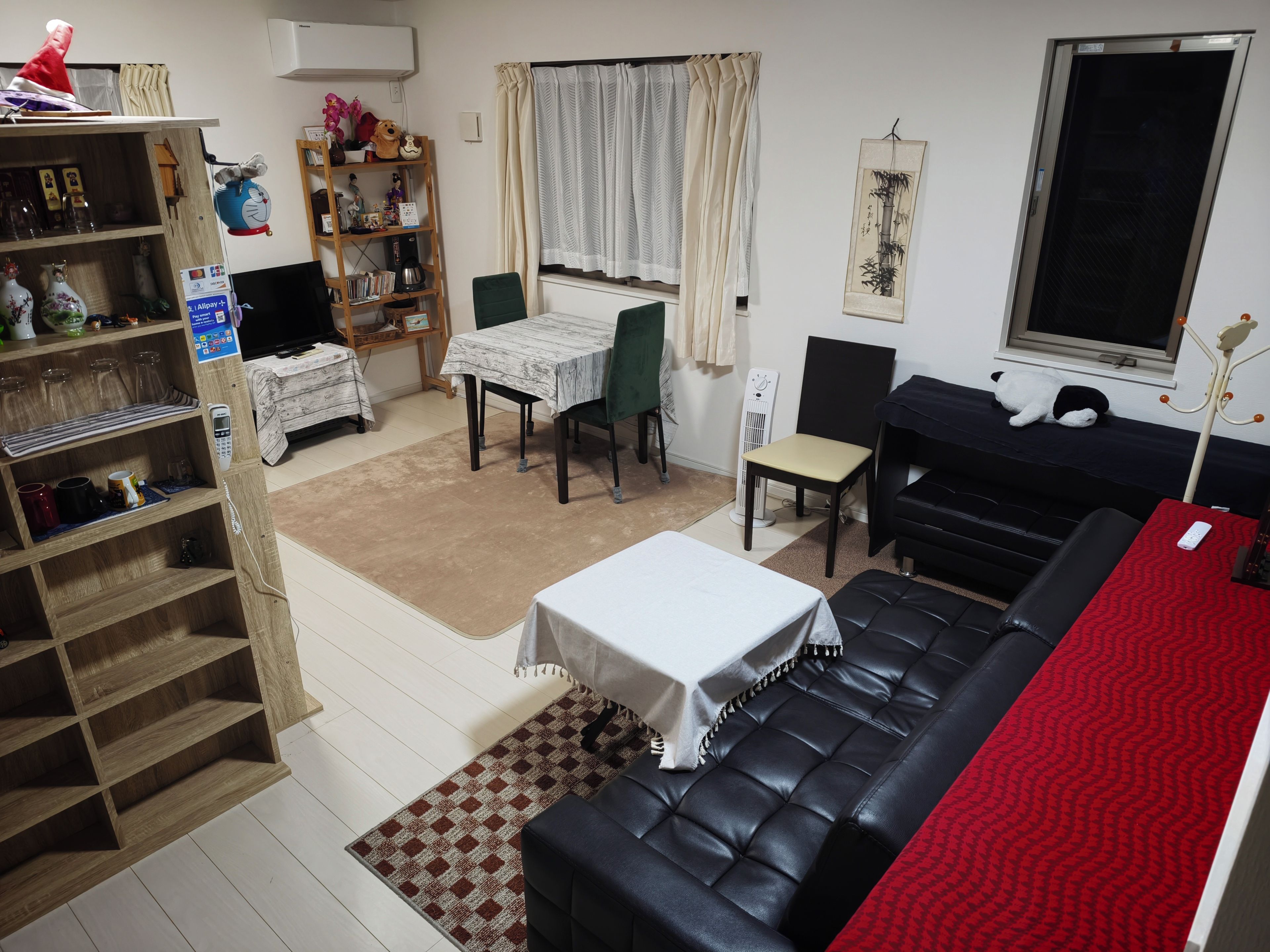 A living room with a black leather sofa, a dining table with chairs, a bookshelf, a small table with a white tablecloth, and a window with curtains.