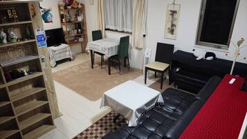 A living room with a black leather sofa, a dining table with chairs, a bookshelf, a small table with a white tablecloth, and a window with curtains.