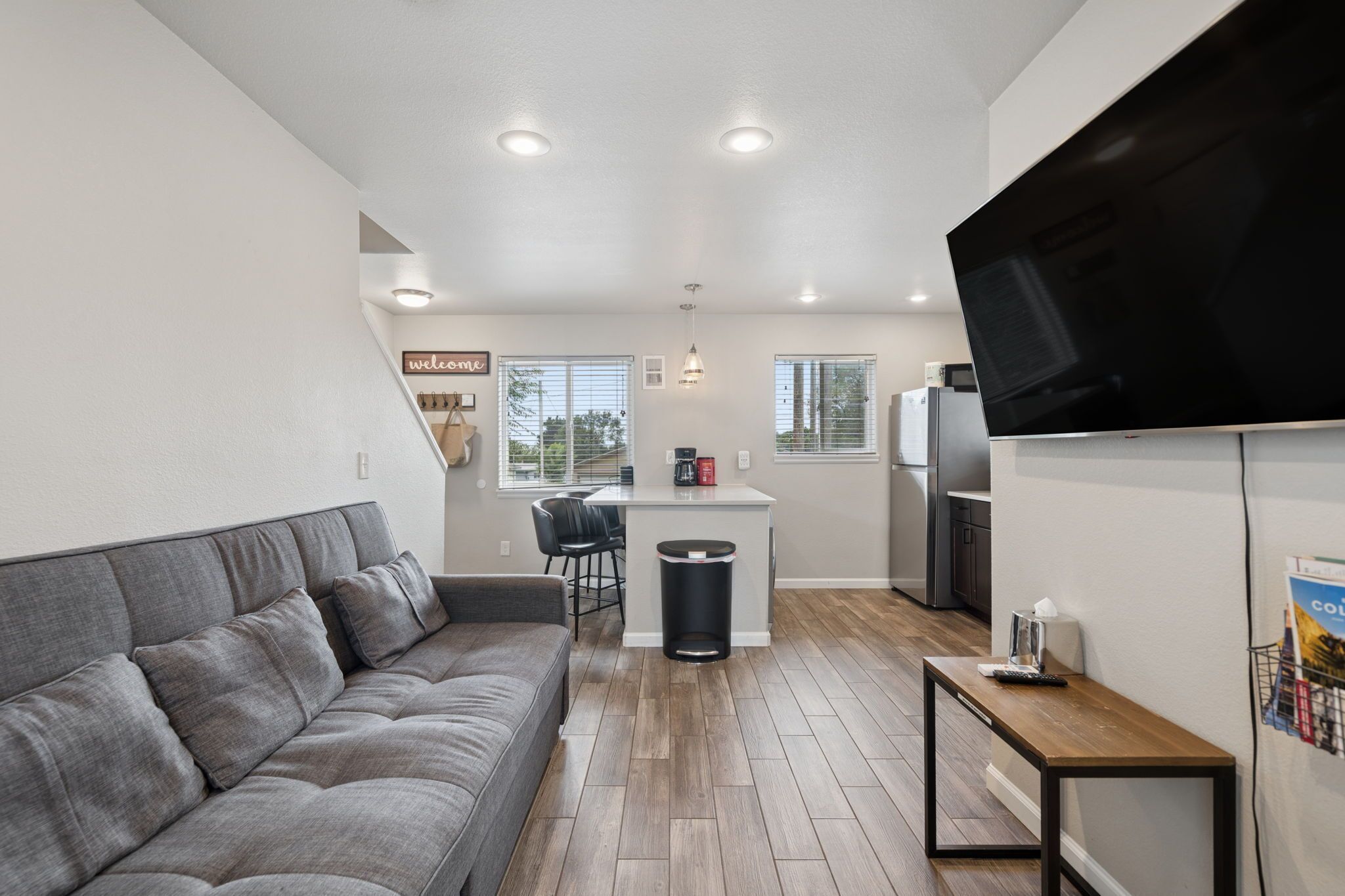 A compact living space with a grey sofa, a small kitchen area, and a flat-screen TV mounted on the wall.