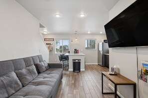 Un espace de vie compact comprenant un canapé gris, un petit coin cuisine et un téléviseur à écran plat fixé au mur.