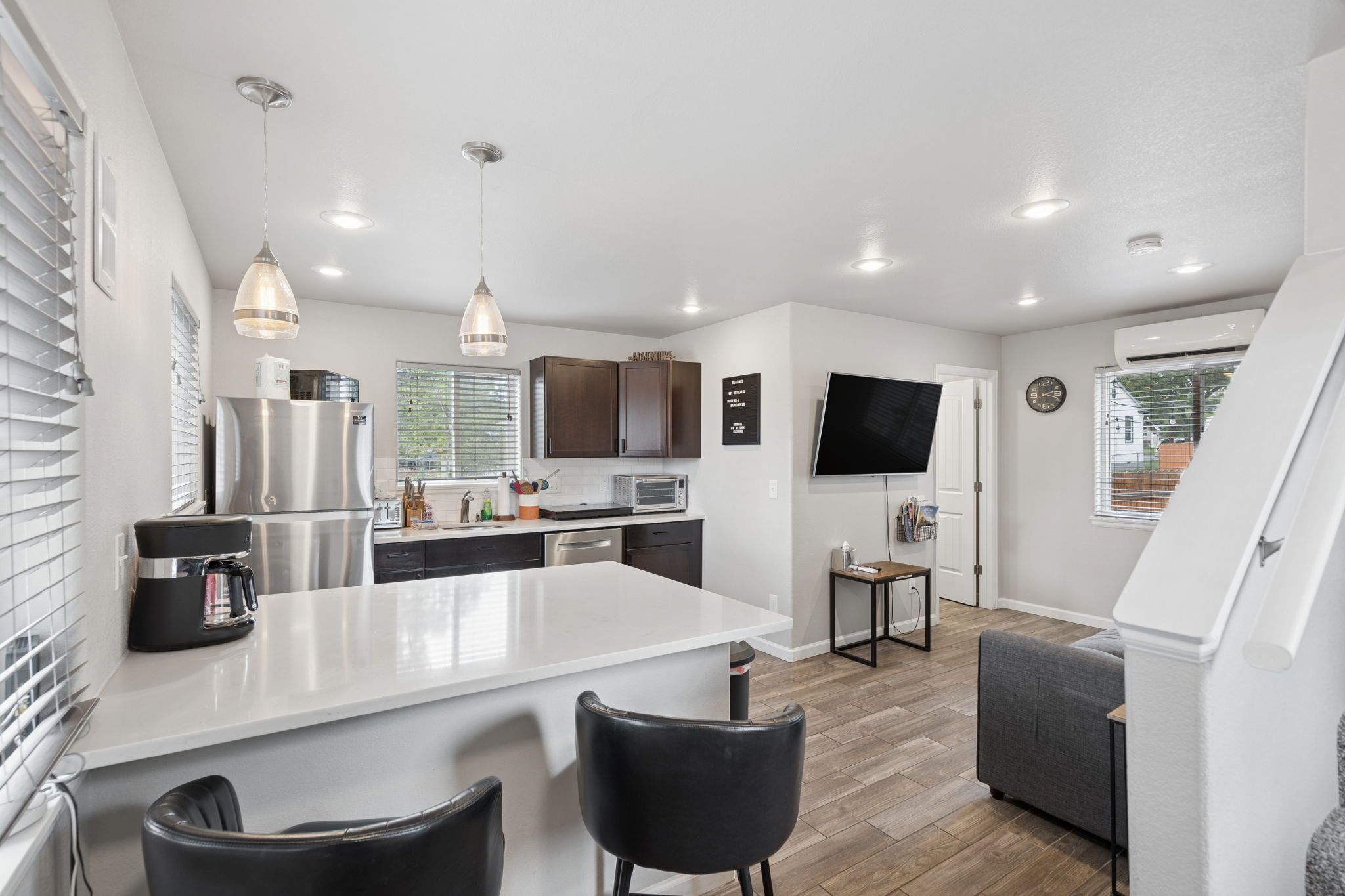 A modern kitchen with a central island, black chairs, a flat-screen TV, and a dining area with a grey sofa.