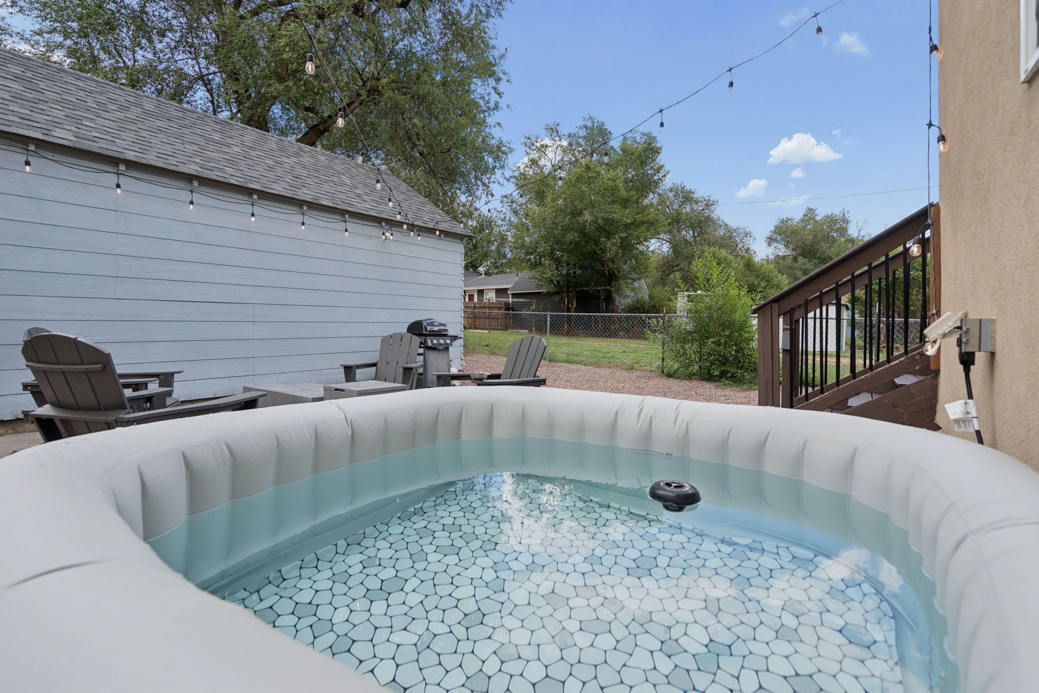 Un jacuzzi al aire libre con asientos y una parrilla, rodeado por un patio trasero con valla y árboles.