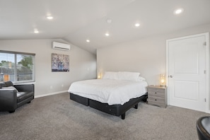 Une chambre à coucher comprenant un lit, une table de chevet, une commode, une fenêtre avec des stores et un appareil de climatisation.