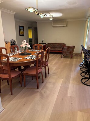 A dining area with a wooden table, chairs, and a patterned table runner. There is a sofa and a fan in the background. - 4 BEDROOM HOUSE IN CECIL HILLS NSW (Cecil Hills)