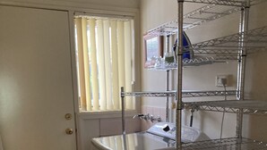Une cuisine compacte équipée d’un lave-linge intégré, d’une étagère métallique et d’une fenêtre avec des stores.