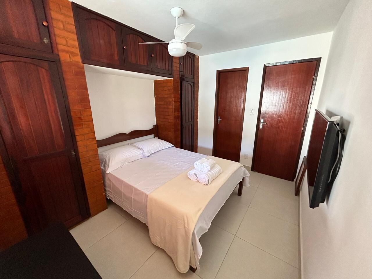 Une chambre à coucher comprenant un lit, une armoire en bois, un ventilateur de plafond et un téléviseur fixé au mur.