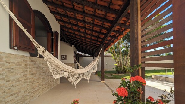 A covered outdoor area with a hammock, a stone wall, and a garden with red flowers. - House 6 minutes walk from Point de Itaúna - Saquarema (Saquarema)