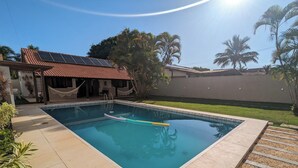 A swimming pool with a tiled deck, a hammock, and a house with solar panels. - House 6 minutes walk from Point de Itaúna - Saquarema (Saquarema)