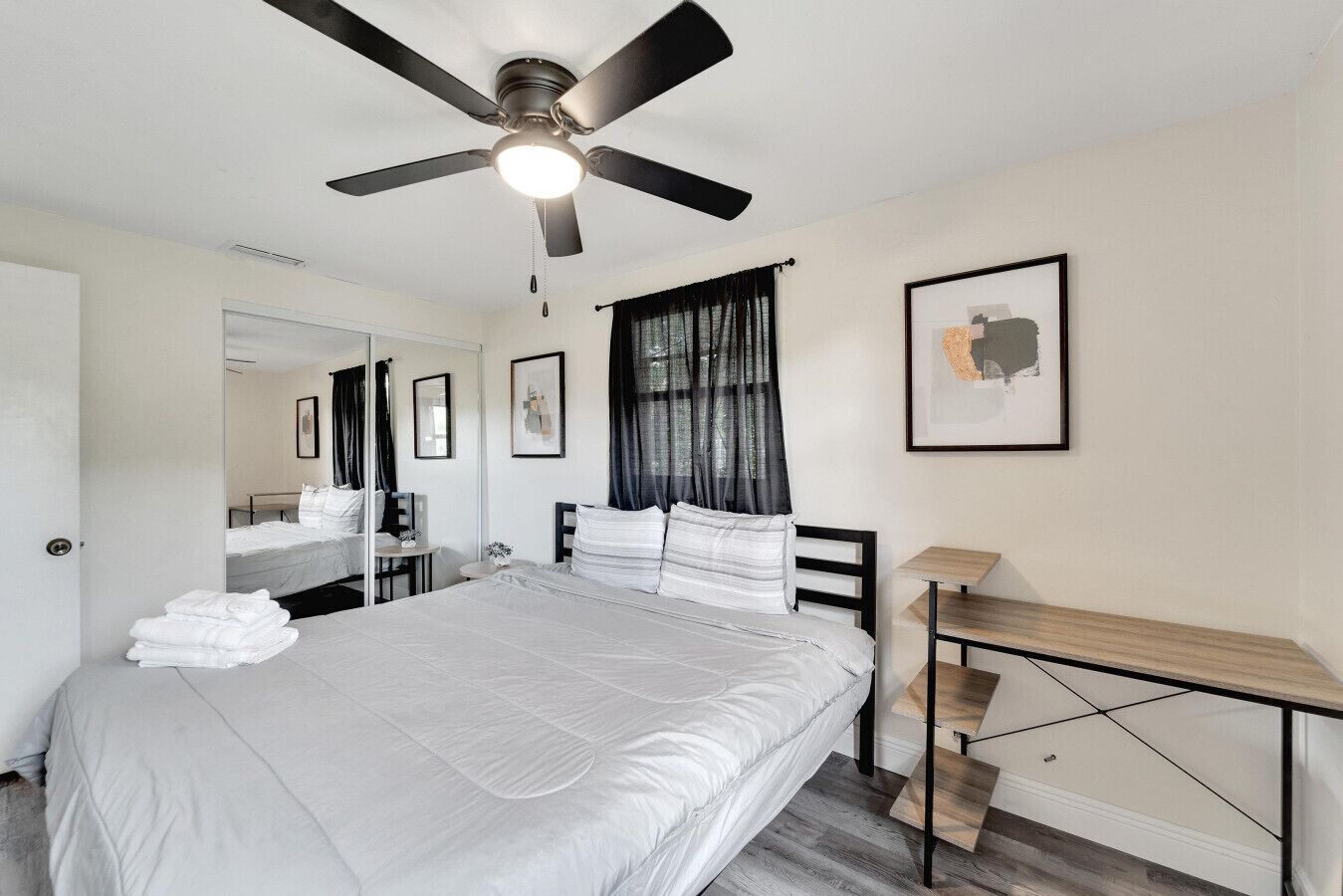 Une chambre à coucher avec un lit, un bureau, un ventilateur de plafond, un miroir et un tableau encadré accroché au mur.