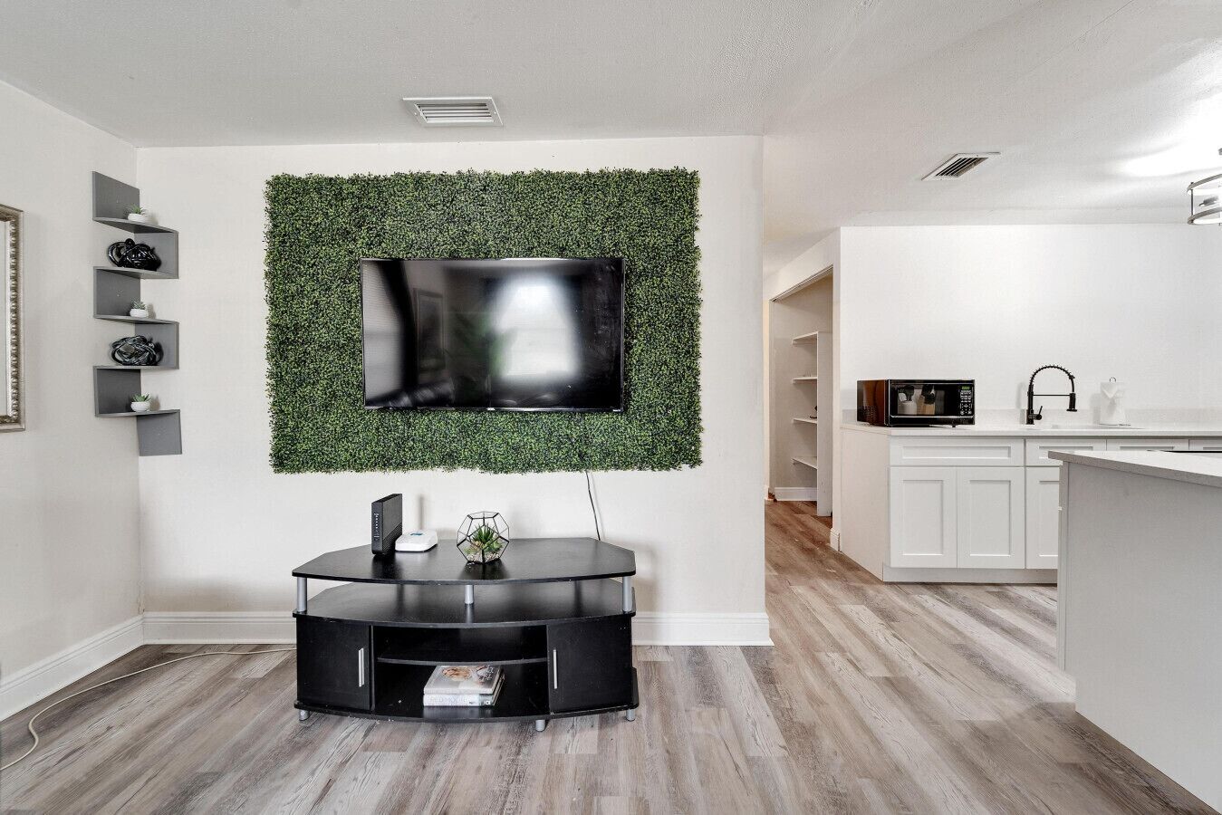 A modern living room with a flat-screen TV mounted on a green wall, a black entertainment unit, and a kitchen area with white cabinets.