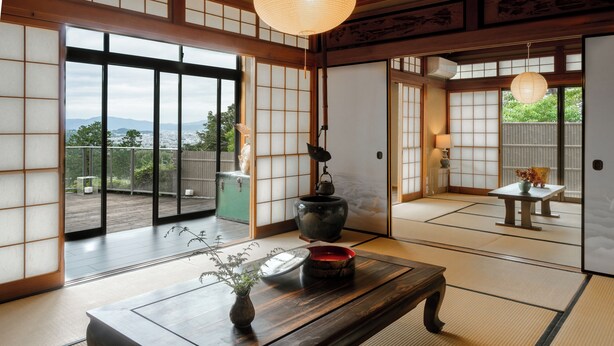 Una tradizionale stanza giapponese con pavimento in tatami, un tavolo in legno con un vaso e un teiera, e porte scorrevoli che danno su un balcone con vista sulle montagne.