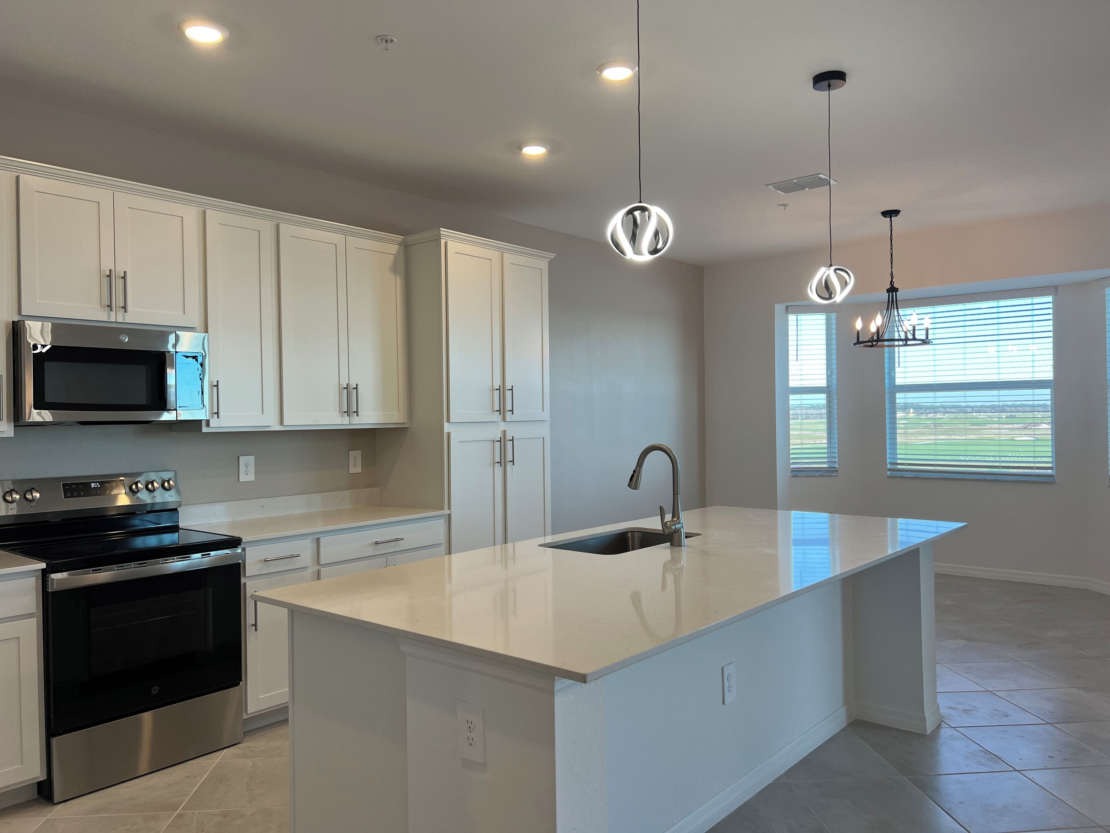 A modern kitchen with white cabinetry, stainless steel appliances, a large island with a sink, pendant lighting, and a view through a window.