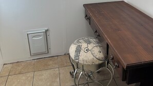 Ein Eckzimmer mit Schreibtisch aus Holz, einem Stuhl im Vintage-Stil, einem Fenster mit Blick auf Bäume und einer weißen Wand mit Thermostat.