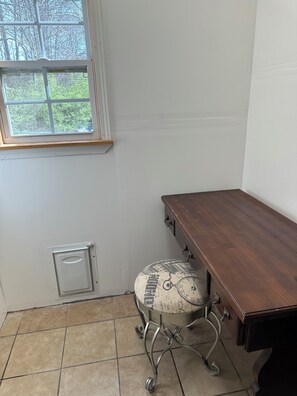 Ein Eckzimmer mit Schreibtisch aus Holz, einem Stuhl im Vintage-Stil, einem Fenster mit Blick auf Bäume und einer weißen Wand mit Thermostat.
