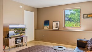A living room with a flat-screen TV, a sofa, a coffee table, and a window with a view of greenery. - The Crag: On the Acadia National Park Border - Deck, Fire Pit, Steps to Trails (Mount Desert)