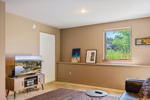 A living room with a flat-screen TV, a sofa, a coffee table, and a window with a view of greenery. - The Crag: On the Acadia National Park Border - Deck, Fire Pit, Steps to Trails (Mount Desert)