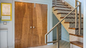 Una puerta de madera con dos pomos, una escalera de madera con escalones y barandillas de metal, y una pared blanca con un letrero amarillo.