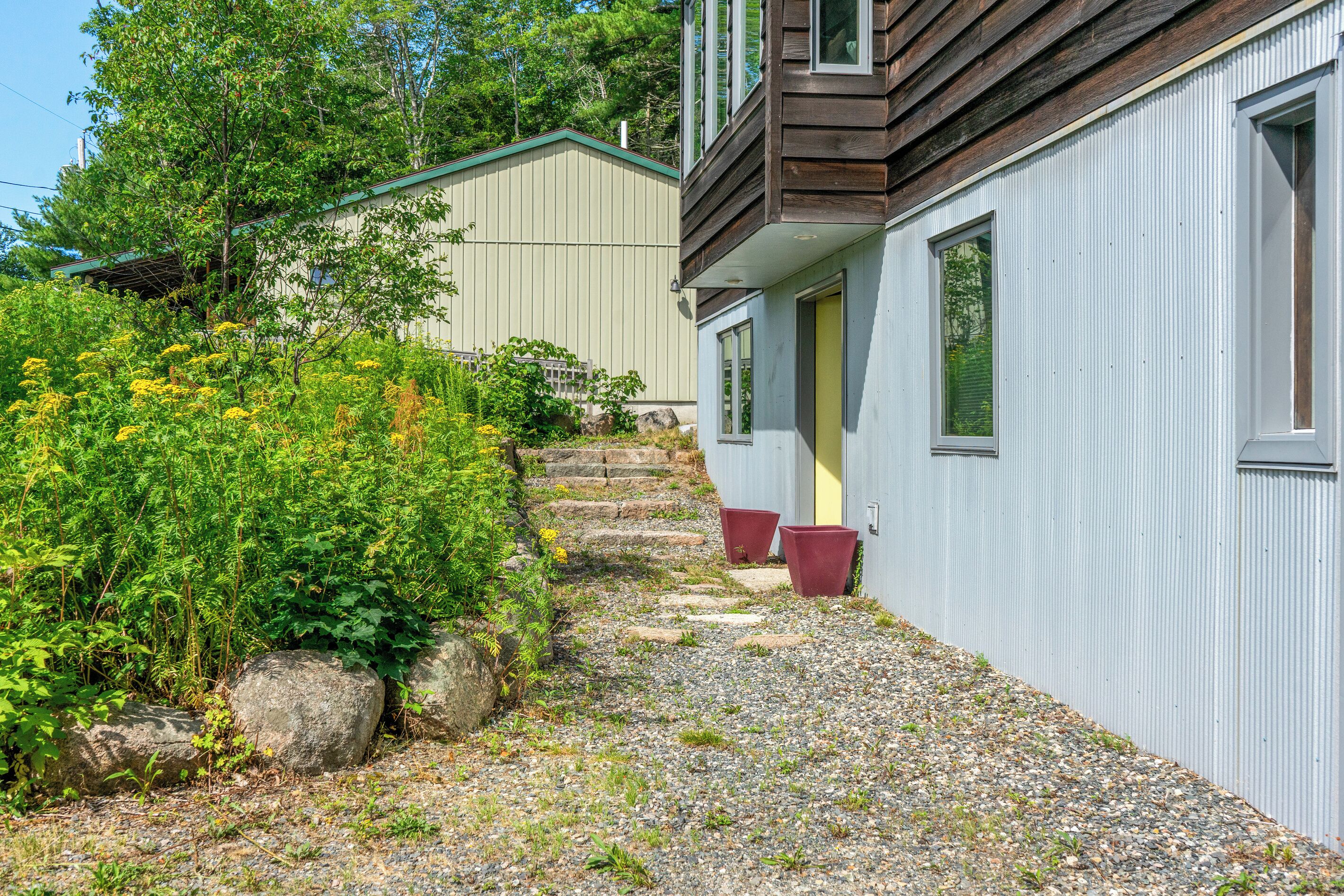 A pathway leading to a building with a wooden facade and a grey exterior. There are steps and potted plants along the path.