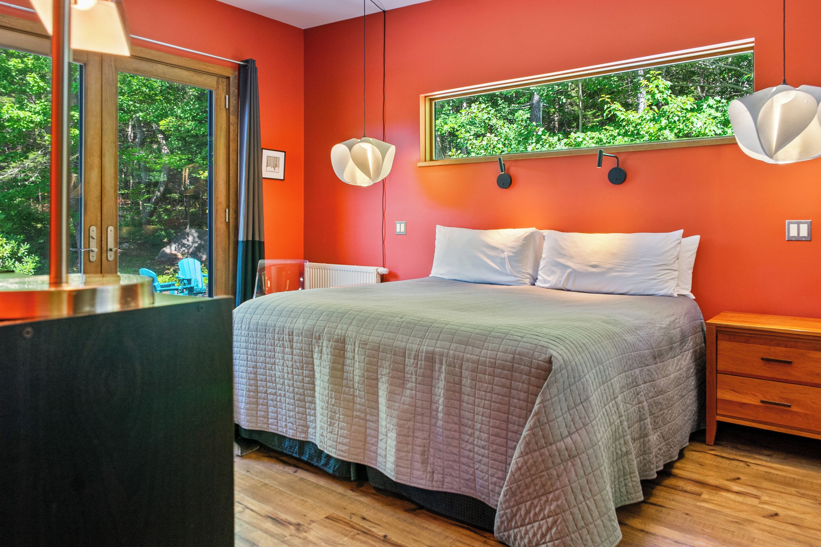 Een slaapkamer met een bed, een nachtkastje, een raam met uitzicht op groen en een wandlamp.