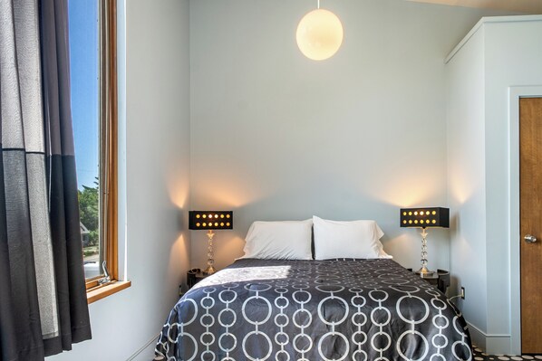A bedroom with a bed, two bedside lamps, a hanging light fixture, and a window with curtains.
