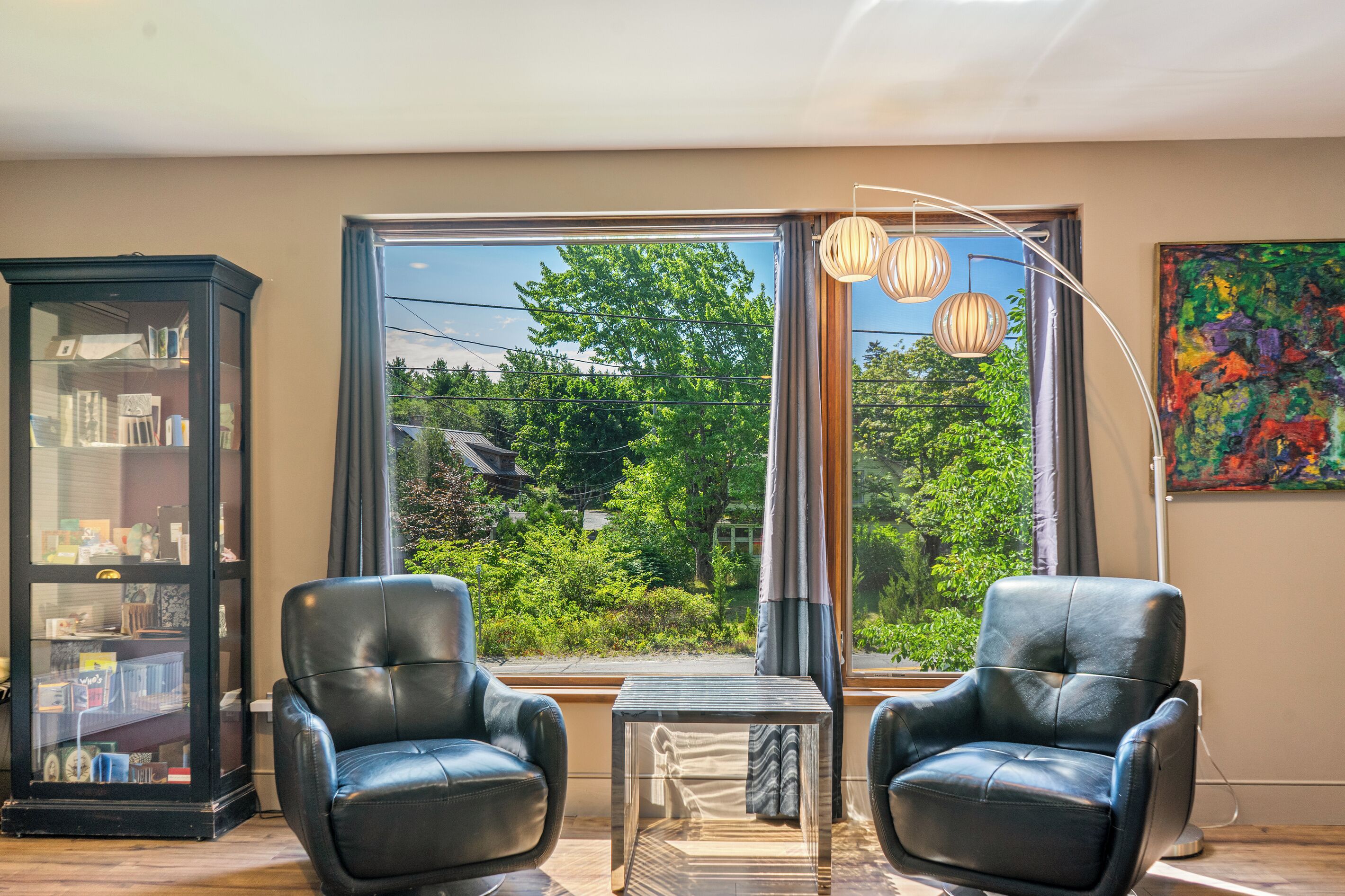 A room with two leather chairs, a small table, a bookshelf, and a large window with a view of greenery.