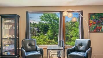 A room with two leather chairs, a small table, a bookshelf, and a large window with a view of greenery.