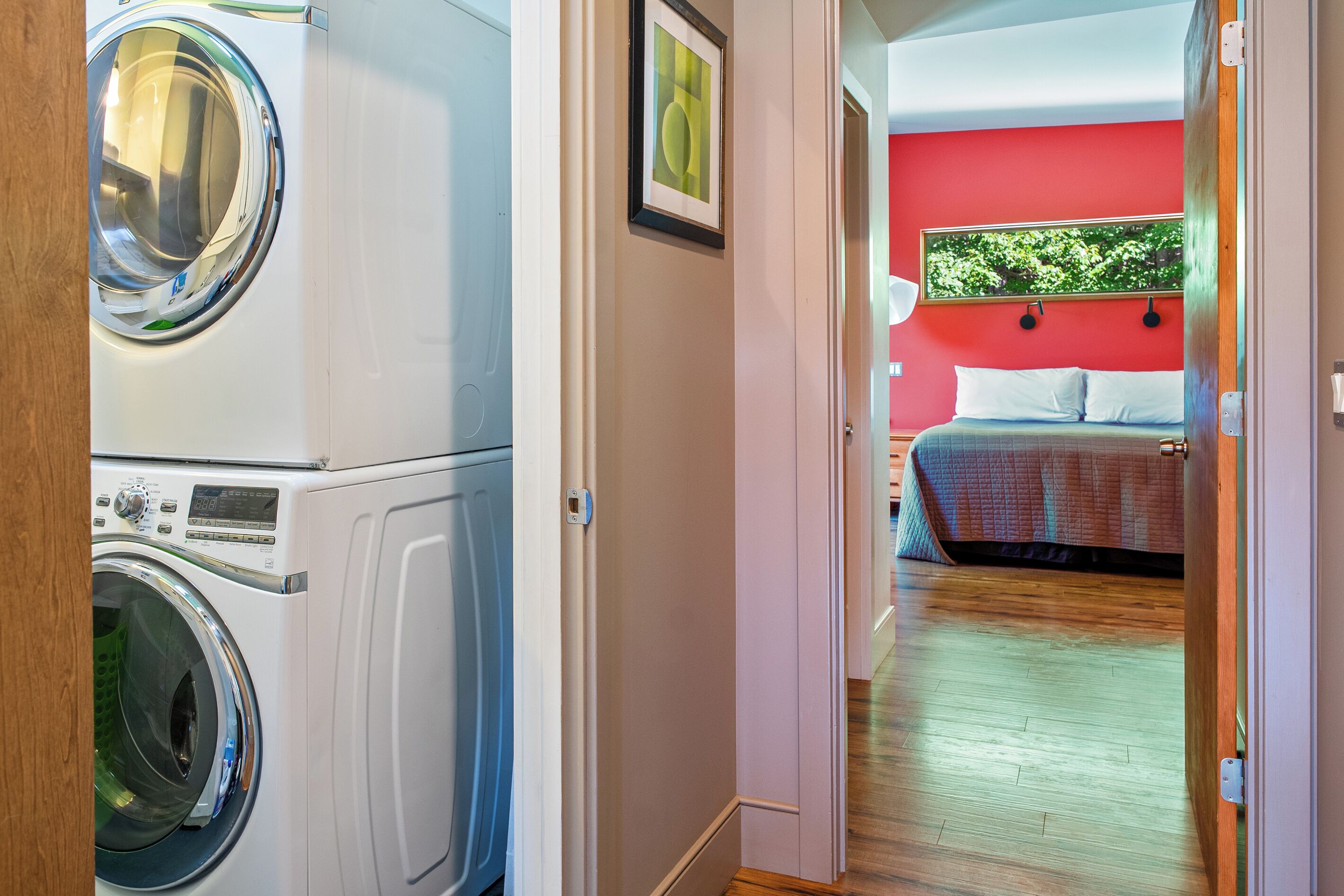 A laundry room with stacked washing machines and a bedroom with a bed and a red wall.