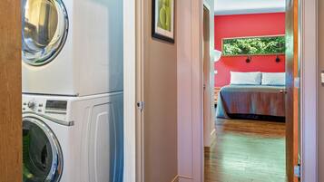 A laundry room with stacked washing machines and a bedroom with a bed and a red wall.